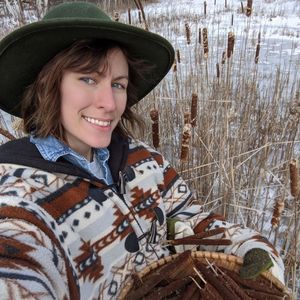 A photo of Val Elkhorn wearing a green hat, holding a basket of cattails in a snow covered marsh.
