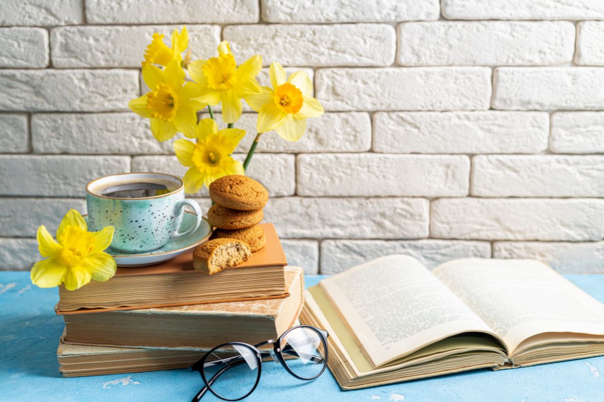 a stack of books with a cup of tea and some cookies sitting on top. Fresh Daffodils sit in the background and an open books sits to the right of the stack.